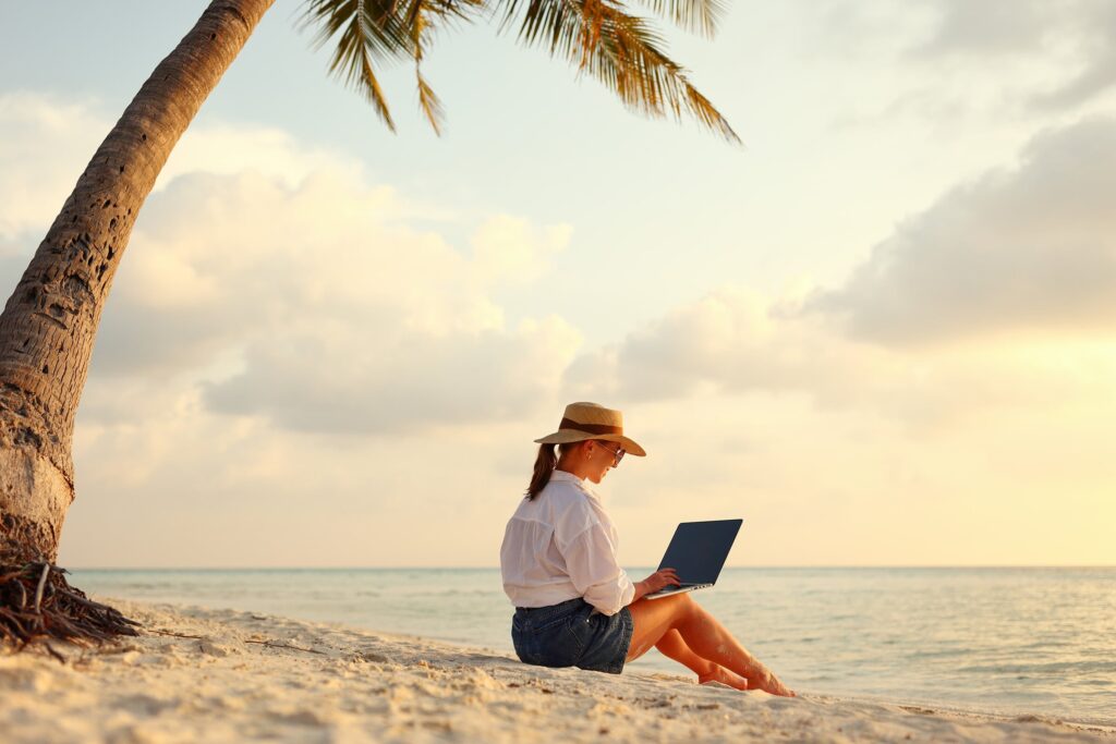 Female travel advisor on a beach under a palm tree, on a laptop looking at platforms for travel agent websites